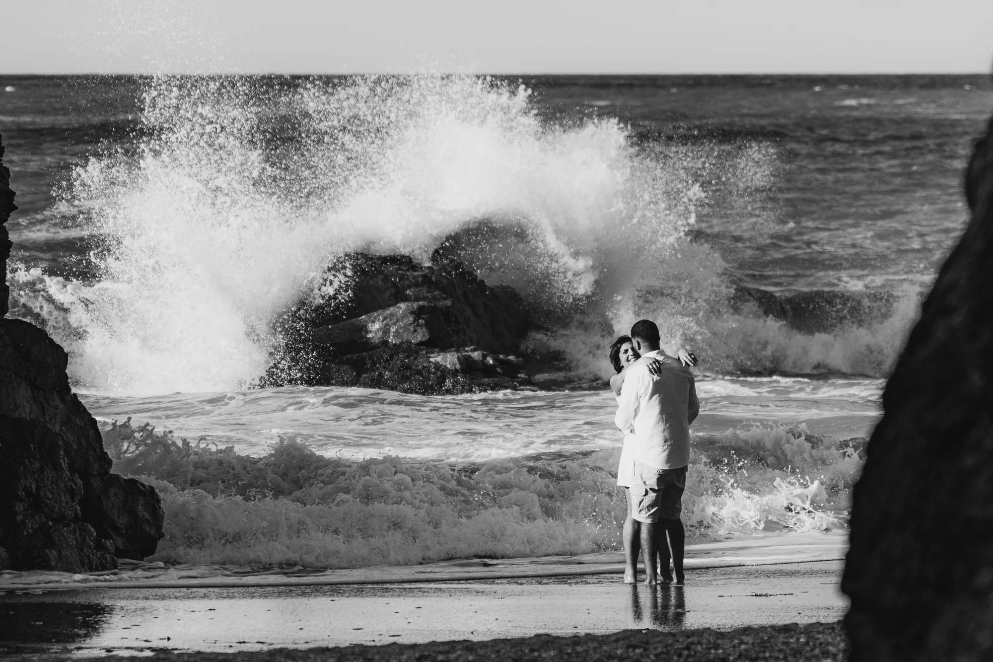 Ola rompiendo junto a pareja de novios en la playa, blanco y negro — Luréa Studio